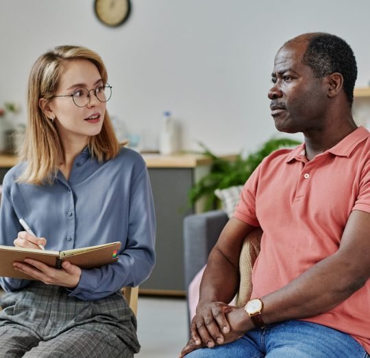 Young psychologist asking questions to patient and making notes in notepad during psychotherapy session