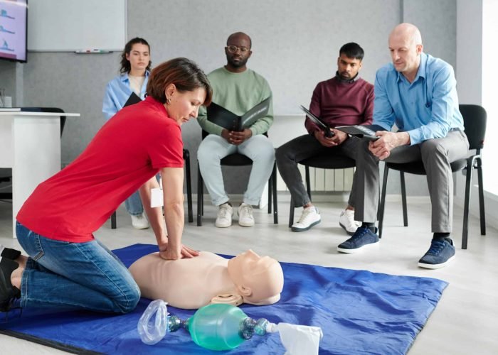 Medical instructor showing CPR on training mannequin on floor to people who sitting on chairs and watching during lesson