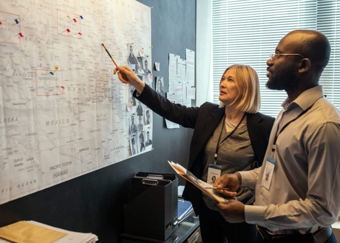 Two FBI agents looking at map on board while mature female pointing at it and making suggestions about possible location of criminal