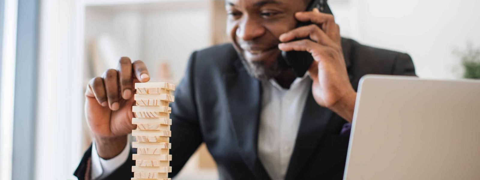 Happy multicultural man in business suit playing jenga while talking on smartphone at office desk with laptop. Cheerful entrepreneur reducing stress at work by enjoying board game during conversation.