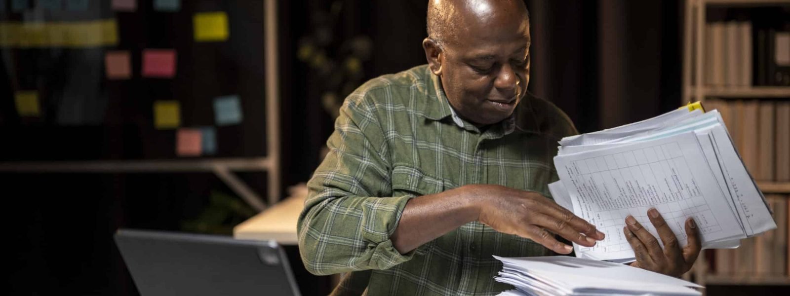 A man is sitting at a desk with a stack of papers in front of him. He is looking at the papers and he is focused on them