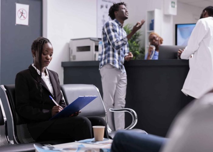 Woman writing medical report papers and checkup form to receive healthcare insurance support before appointment with physician. Patient sitting in waiting room area at hospital facility lobby.