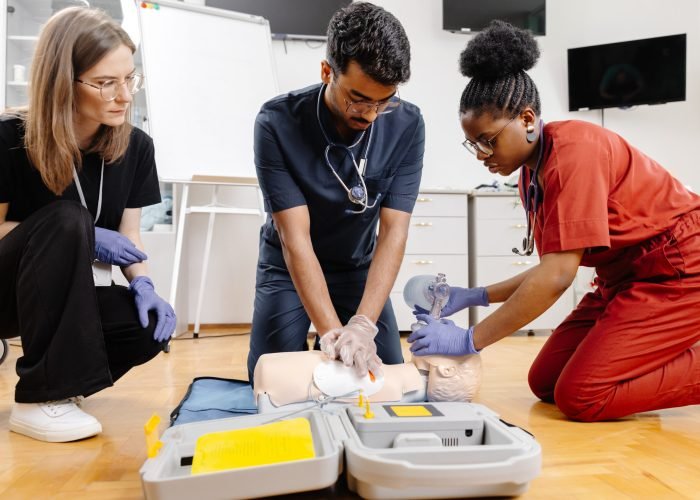 Three individuals practice CPR techniques on a training dummy in a classroom setting.