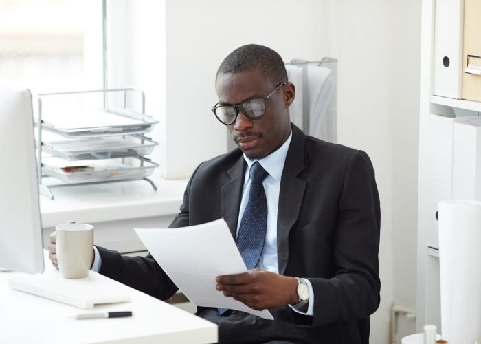 Serious African businessman in eyeglasses and in formal wear sitting at the table examining the document and holding cup of coffee at office