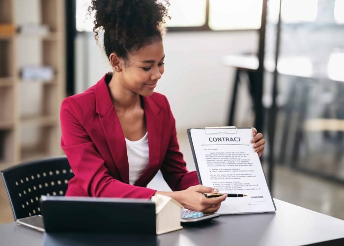 African american businesswoman in suit showing house model and pointing on contract document to presentation about renting or buying and insurance housing while working on tablet in workspace modern.